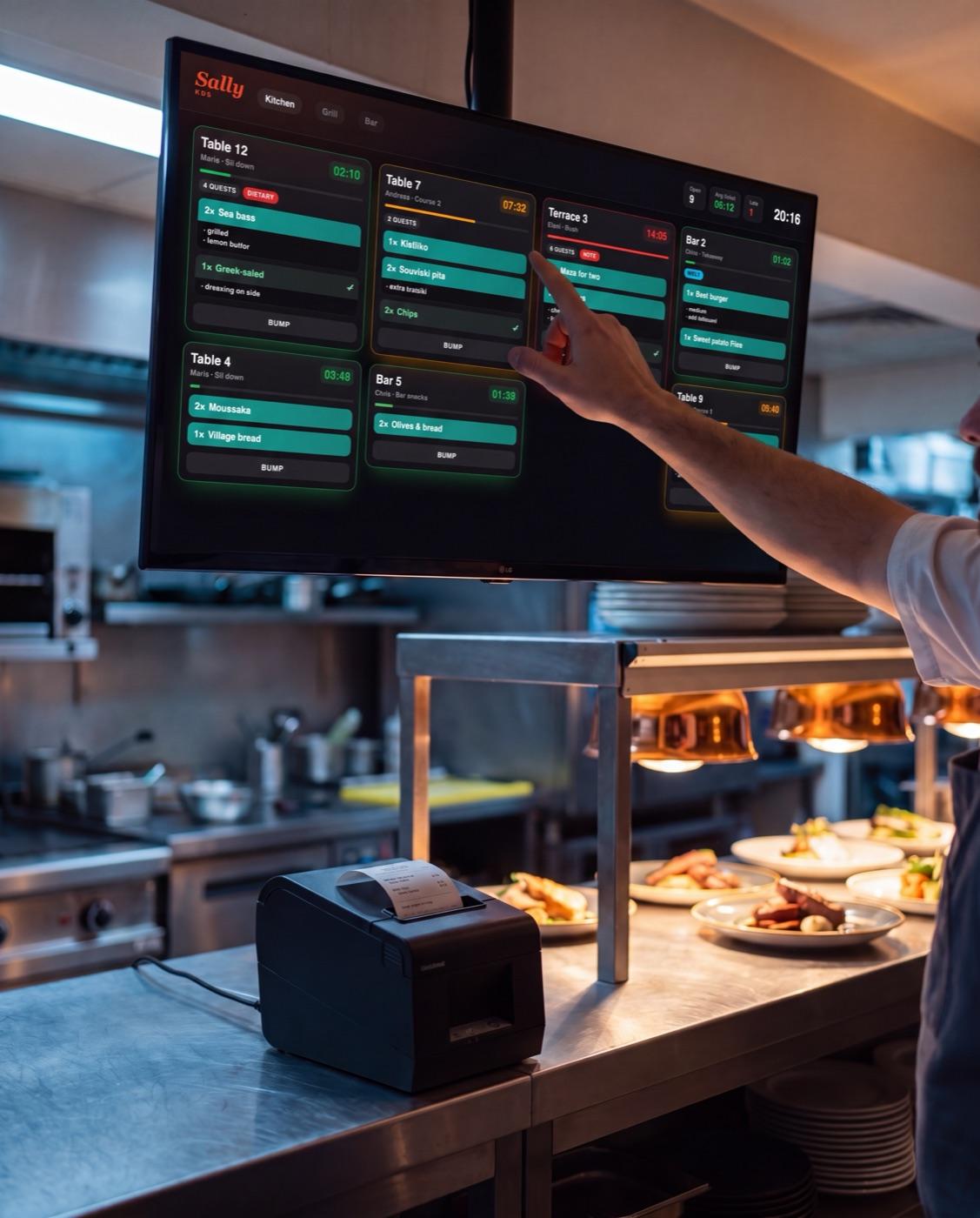 A chef tapping an order on the Sally kitchen display above the pass, next to a ticket printer
